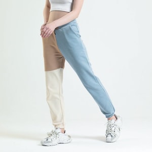 May include: A woman wearing a white tank top and colour-blocked trousers. The trousers are beige, cream, and light blue. She is wearing white trainers.