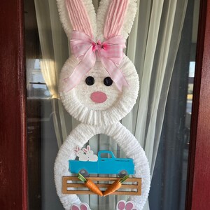 May include: A white Easter bunny wreath with pink accents. The bunny has pink ears, a pink bow, and a pink nose. A blue truck carries two white bunnies. Two orange carrots and pink paw prints complete the design.