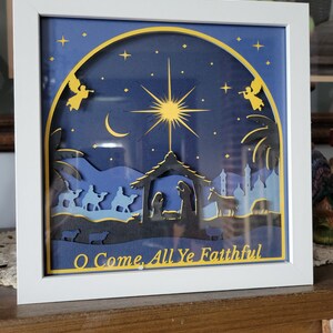 Könnte beinhalten: Gerahmtes Papierkunstwerk mit einer Krippenszene. Das Kunstwerk zeigt einen Stern, Engel und den Text "O Come, All Ye Faithful". Der Rahmen ist weiß, und der Hintergrund ist dunkelblau mit goldenen Akzenten.