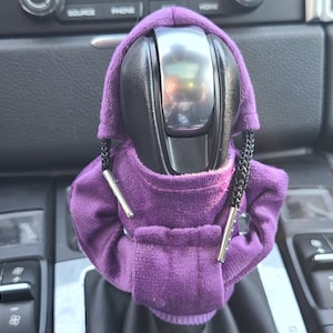 May include: Purple hooded gear shifter cover with black strings and silver accents. The cover is made of soft, plush fabric and fits over a standard gear shifter.
