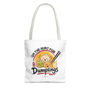 Könnte beinhalten: Eine weiße Tragetasche mit einer grafischen Illustration von drei lächelnden Teigtaschen in Grün, Orange und Beige. Das Design enthält den schwarzen Schriftzug ON THE HUNT FOR Dumplings, ein Paar schwarze Essstäbchen und einen kleinen roten Stempel mit asiatischen Schriftzeichen.