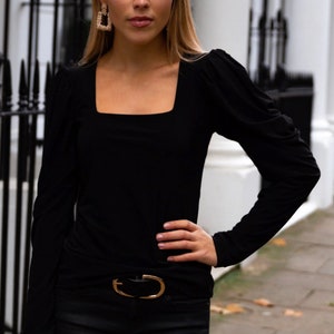 Pode incluir: Uma mulher vestindo um top preto de manga comprida com decote quadrado e mangas bufantes. Ela está usando um cinto dourado e calças pretas. Ela está segurando uma clutch dourada.