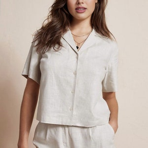 May include: A woman wearing a light beige linen shirt with a collared neckline and short sleeves. The shirt has a button-up front and is paired with matching linen shorts. The shorts have a relaxed fit and feature cargo pockets.
