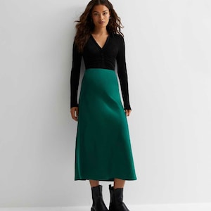 Könnte beinhalten: Eine Frau trägt ein schwarzes Langarmshirt und einen grünen Satin-Midirock. Sie trägt schwarze Stiefeletten.