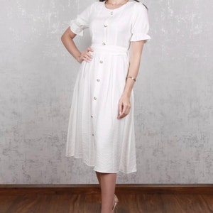 May include: A white midi dress with short sleeves and a button-up front. The dress has a belt at the waist and a slightly flared skirt.