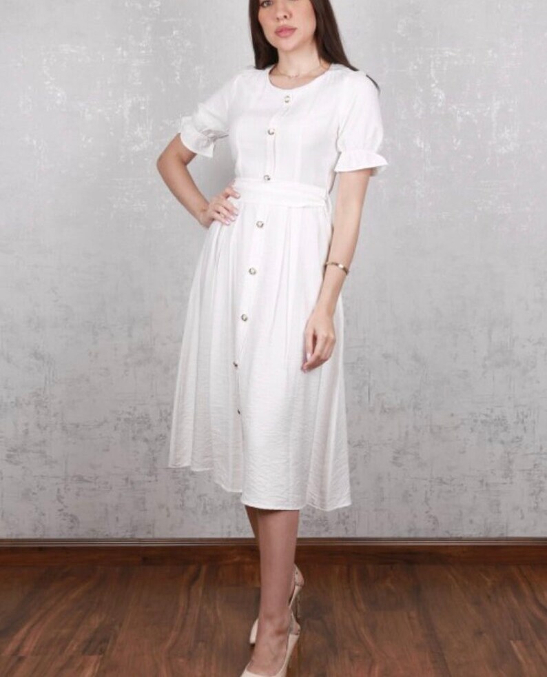 May include: A white midi dress with short sleeves and a button-up front. The dress has a belt at the waist and a slightly flared skirt.