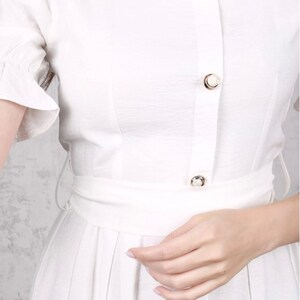 May include: A white button-down dress with a ruffled short sleeve and a cinched waist. The dress has a row of pearl buttons down the front.