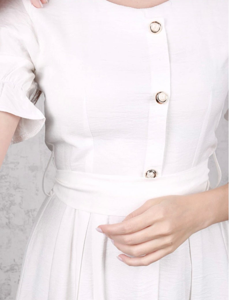 May include: A white button-down dress with a ruffled short sleeve and a cinched waist. The dress has a row of pearl buttons down the front.