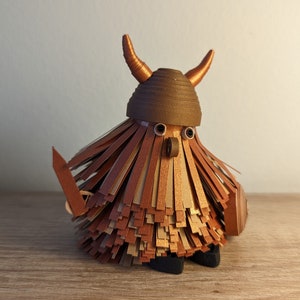 May include: A brown papercraft figurine of a warrior with a horned helmet, a sword, and a shield. The figurine is made of layered paper strips in shades of brown, gold, and beige.