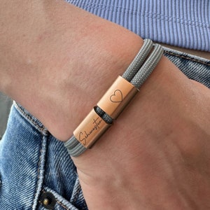 Könnte beinhalten: Ein graues Seil-Armband mit einer roségoldenen Stange, die mit dem Wort "Schwester" und einem Herzen graviert ist.