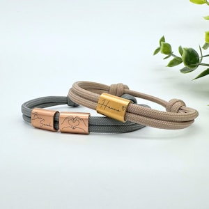 Könnte beinhalten: Zwei Freundschaftsarmbänder mit personalisierten, gravierten Metallperlen. Ein Armband ist grau mit den Namen "Ben Sarah" und einem Herzsymbol. Das andere Armband ist beige mit dem Namen "Hanna" und einem Herzsymbol.