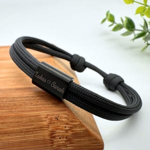 Könnte beinhalten: Ein schwarzes Seil-Armband mit einem schwarzen Metallverschluss, der mit dem Text "Lukas ♥ Sarah" graviert ist.