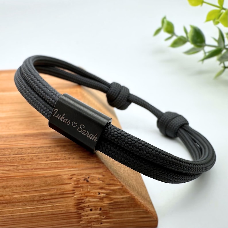 Dunkelgraues Armband das einen schwarzen Edelstahlschieber besitzt. Auf dem Schieber ist der Name Lukas ein Herz und Sarah hintereinander graviert.
