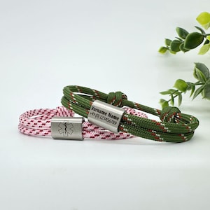 Peut inclure: Deux bracelets en paracorde avec des étiquettes en métal. Un bracelet est vert avec des rayures rouges et blanches et porte une étiquette gravée avec "Vorname Name +49 (0) 123456789". L'autre bracelet est rose avec des rayures rouges et blanches et porte une étiquette avec un symbole médical.