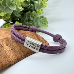 Op de afbeelding: Een paarse paracord armband met twee strengen en een zilveren metalen tag met de coördinaten "52°30'58.25"N 13°22'36.96"O" erop gegraveerd.