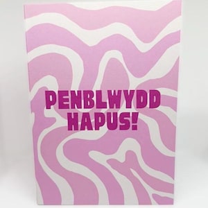 Puede incluir: Tarjeta de felicitación rectangular con un patrón ondulado rosa y blanco. El texto "PENBLWYDD HAPUS!" se muestra en negrita, en color rosa oscuro. La tarjeta está diseñada para mantenerse de pie.