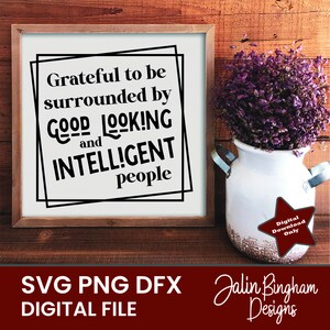 May include: A white framed sign with black text that reads "Grateful to be surrounded by GOOD LOOKING and INTELLIGENT people". The sign is sitting on a wooden surface with a purple flower arrangement in a white pitcher.