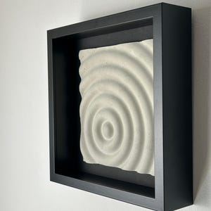 May include: A white, textured, circular design framed in a black frame. The design is raised and appears to be made of concrete.