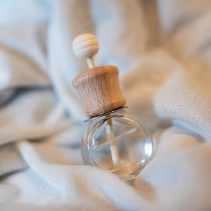 May include: A clear glass bottle with a wooden lid and a white diffuser stick. The bottle is filled with a clear liquid and is sitting on a white fabric surface.