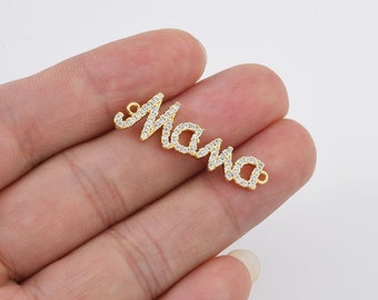 Puede incluir: Un dije dorado con la palabra "Mama" escrita en cursiva. Las letras están incrustadas con pequeños cristales transparentes. El dije tiene pequeños bucles en cada extremo para sujetarlo a un collar o pulsera. El dije está en la mano de una persona.