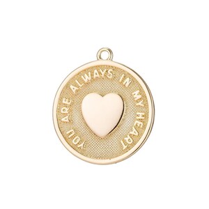 Puede incluir: Colgante dorado con diseño de corazón. El colgante tiene forma circular con las palabras "YOU ARE ALWAYS IN MY HEART" grabadas alrededor del borde. Una forma de corazón está en el centro del colgante.