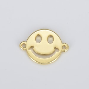 May include: A gold-colored smiley face charm with a circular shape. The charm features a cut-out smiling mouth and two eye holes. Small loops are attached on either side of the charm for stringing or attaching to other jewelry pieces. The charm is set against a white background.