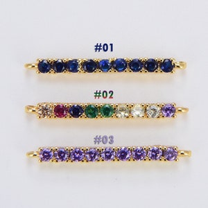 以下が含まれることがあります： 異なる色のクリスタルが付いた、金色のバーチャームが3つあります。最初のチャームは青いクリスタル、2番目のチャームは赤、青、緑、白のクリスタル、3番目のチャームは紫のクリスタルが付いています。チャームには#01、#02、#03のラベルが付いています。