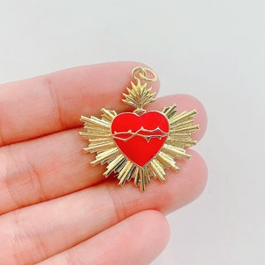 以下が含まれることがあります： 赤いハートに茨の冠をあしらった、太陽光線模様のゴールドカラーのペンダント。ペンダントには、チェーンやネックレスに取り付けるためのループが付いています。デザインは詳細で、色は鮮やかです。