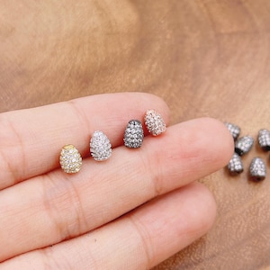 May include: Close-up of several small, oval-shaped earrings. The earrings are encrusted with small, sparkling stones. The earrings are in various colors, including gold, silver, black, and rose gold. Additional earrings are scattered in the background.