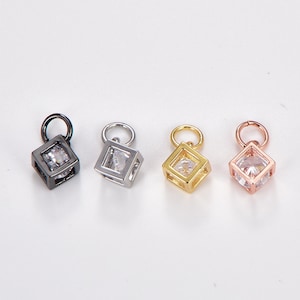 May include: Four cubic charms in black, silver, gold, and rose gold. Each charm features a clear, square-cut gemstone set within a cube frame. The charms have small loops for attaching to jewelry.