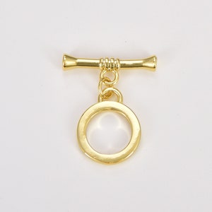 May include: Gold-toned metal toggle clasp with a round ring. The clasp has a bar with a decorative design and a ring attached to the bar.