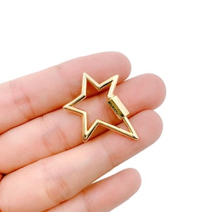 May include: A gold-colored star-shaped clasp with a rectangular locking mechanism. The clasp is held in a hand against a white background. The star has five points and a hollow center.
