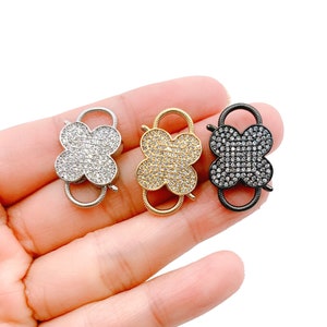 May include: Three clover-shaped clasps in silver, gold, and black. Each clasp features a four-leaf clover design encrusted with small, sparkling crystals. The clasps are held in a hand against a white background.