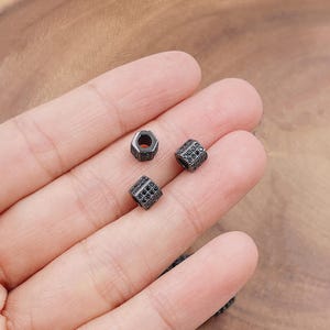 May include: Close-up of three small, dark gray metal beads with intricate detailing. Two are cube-shaped, adorned with small, black stones. The third bead is hexagonal. These beads are likely for jewelry making.