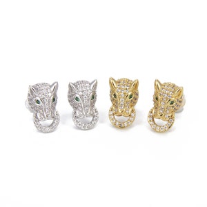 Könnte beinhalten: Ein Paar Ohrringe mit einem Wolfskopf-Design, besetzt mit klaren und grünen Zirkonia-Steinen. Die Ohrringe sind in Silber und Gold erhältlich.