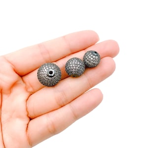 May include: Three black round beads with a pave setting of clear rhinestones. The beads are different sizes and are held in a hand.