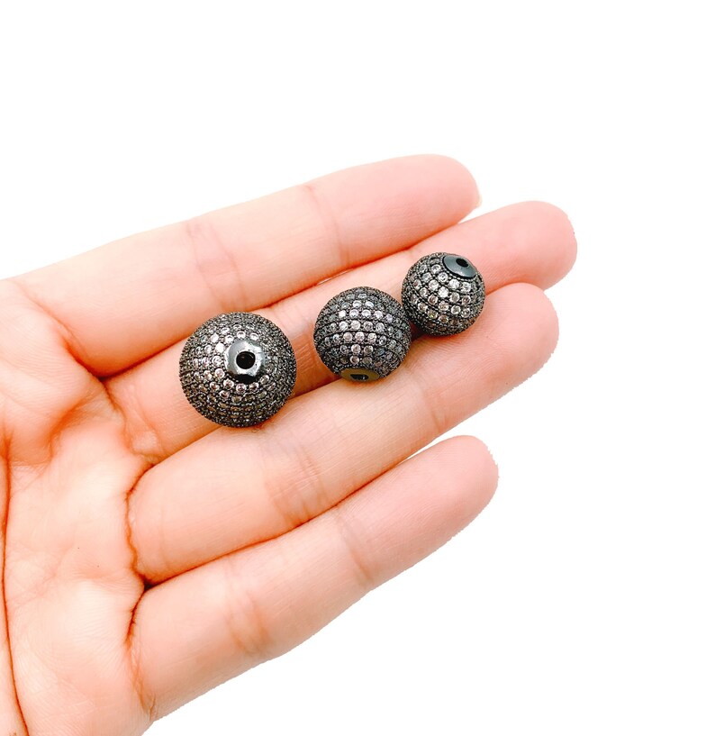 May include: Three black round beads with a pave setting of clear rhinestones. The beads are different sizes and are held in a hand.