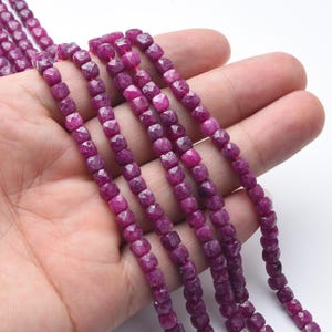 May include: Strands of faceted, deep magenta-colored gemstone beads. The beads are square-shaped with multiple facets, strung together on a thin, pink string. The beads are held in a hand against a white background. Ideal for jewelry making.