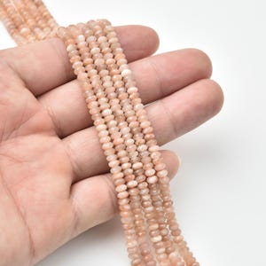 May include: Strands of peach-colored gemstone beads. The beads are small, faceted, and strung together, creating a delicate and textured appearance. The beads are held in a hand against a white background. Ideal for jewelry making.