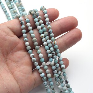 May include: Strands of faceted, round beads in shades of light blue, white, and gray. The beads are held in a hand against a white background. The beads are strung together, ready for jewelry making.