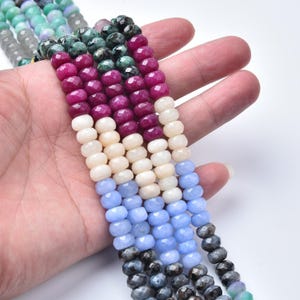 May include: Strands of gemstone beads in various colors, including deep red, cream, light blue, and dark gray. The beads are faceted and strung together, creating a textured and colorful display. The gemstones are held in a hand.