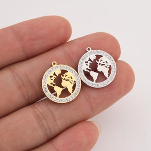 May include: Two round pendants featuring a world map design. One pendant is gold-toned, the other is silver-toned. Both have a border of clear crystals. Each pendant has a small loop at the top for attaching to a chain or other jewelry.