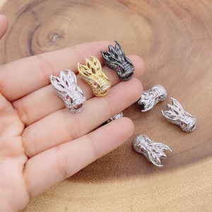 May include: Dragon head beads in silver, gold, and black. Each bead features intricate detailing and a red gemstone eye. The beads are small, perfect for jewelry making, and are displayed on a wooden surface.