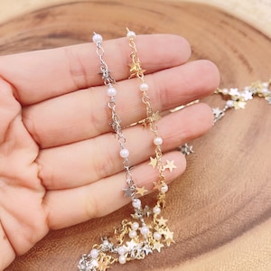 Op de afbeelding: Close-up van twee delicate kettingen. De ene is zilverkleurig, de andere goudkleurig. Beide hebben kleine sterbedels en kleine witte parels geregen aan een ketting. De kettingen worden gepresenteerd tegen een warme, neutrale achtergrond.