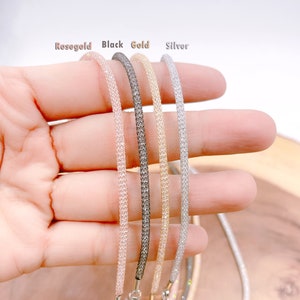 May include: Five different colored chain necklaces, rose gold, black, gold, and silver, are displayed on a hand. The necklaces are made of a fine mesh material.