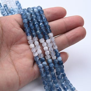 May include: Strands of faceted blue and white gemstone beads. The beads are square-shaped and have a polished finish. The beads are held in a hand, showcasing the color variations and the texture of the gemstones.