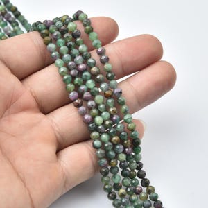 May include: Strands of faceted gemstone beads in shades of green, purple, and brown. The beads are small and round, with a polished, reflective surface. The beads are held in a hand against a white background. The beads are likely for jewelry making.