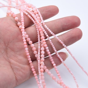 May include: Several strands of pink gemstone beads in varying sizes are held in a hand. The beads range from small seed beads to larger, round beads. The color is a soft, light pink.