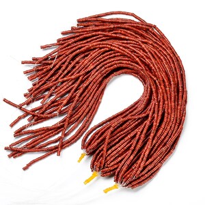 May include: A collection of reddish-brown seed beads, tightly strung together. The beads are small and cylindrical, with a slightly glossy finish. The strands are bundled together, with yellow thread securing the ends. The beads are likely for jewelry making or other crafts.