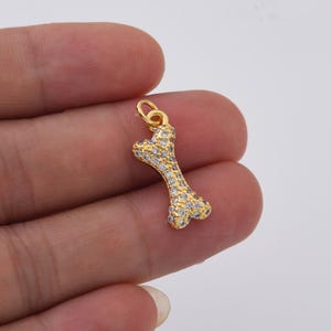 May include: A gold-colored bone-shaped charm, encrusted with small, clear crystals. The charm has a loop at the top for attaching to a necklace or bracelet. The charm is held in a person's hand.